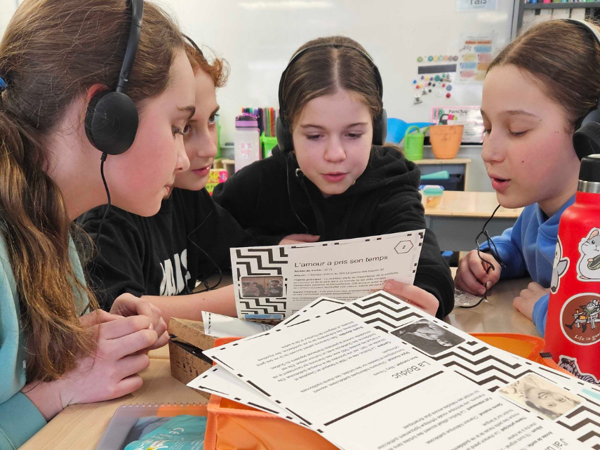 4 élèves avec des casques d'écoute. L'une d'elle lit à haute voix depuis une feuille de papier.